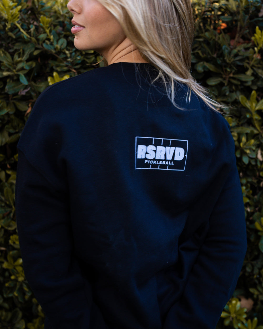 Person wearing a black sweatshirt with a logo against a green hedge background