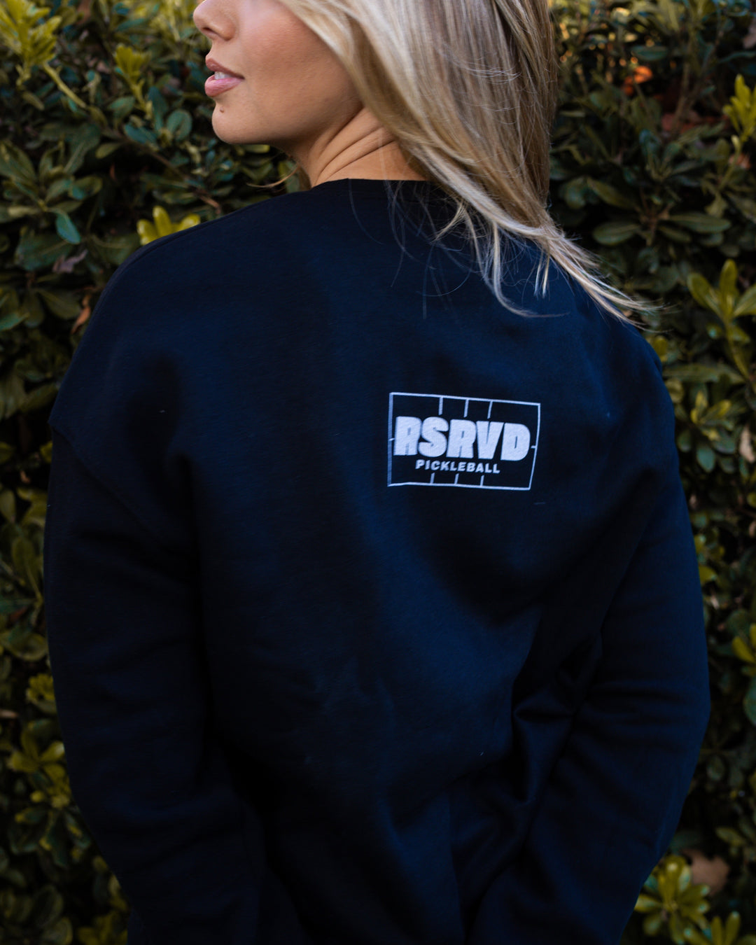 Person wearing a black sweatshirt with a logo against a green hedge background