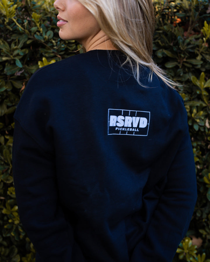 Person wearing a black sweatshirt with a logo against a green hedge background