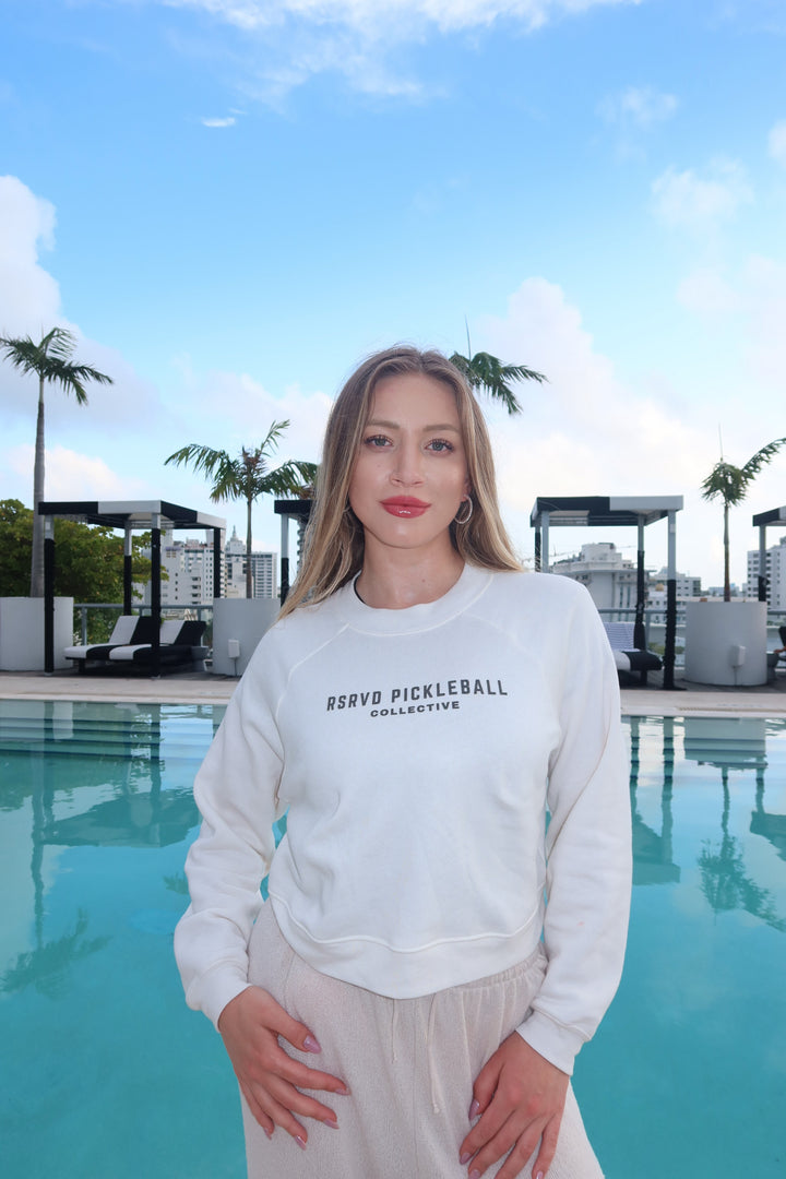 Woman wearing a white sweatshirt with text, standing by a pool with palm trees and buildings in the background.