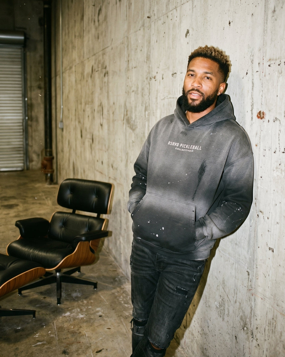 Man wearing a gray hoodie standing in an industrial setting with a chair and concrete wall.