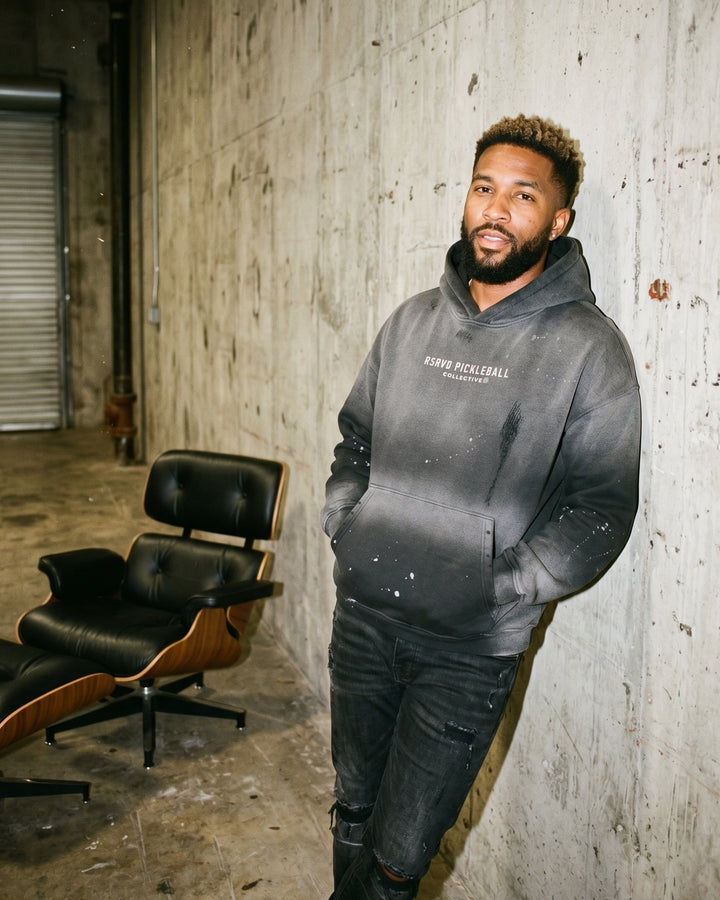 Man wearing a gray hoodie standing in an industrial setting with a chair and concrete wall.