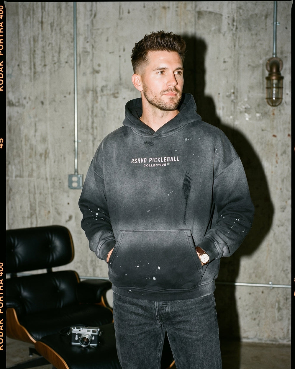 Man wearing a black hoodie with text, standing against a concrete wall.