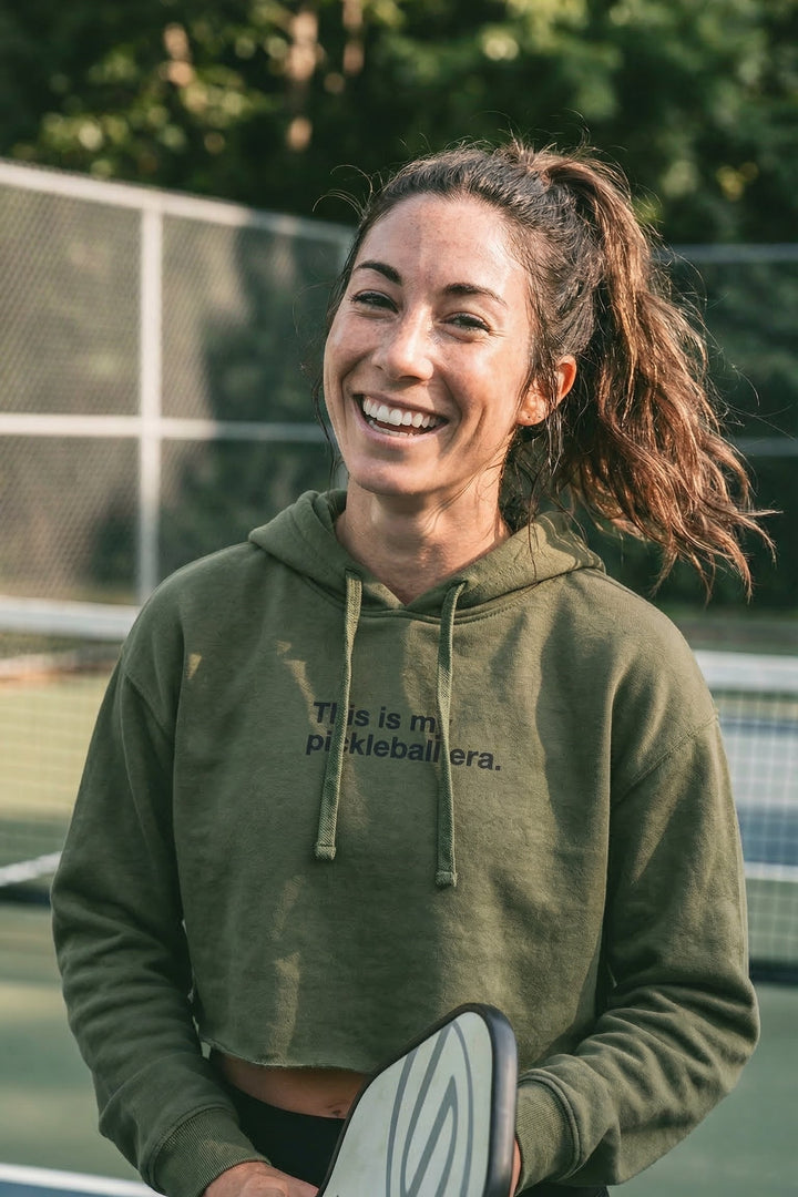 Woman wearing a green hoodie with text, holding a paddle, on a tennis court.