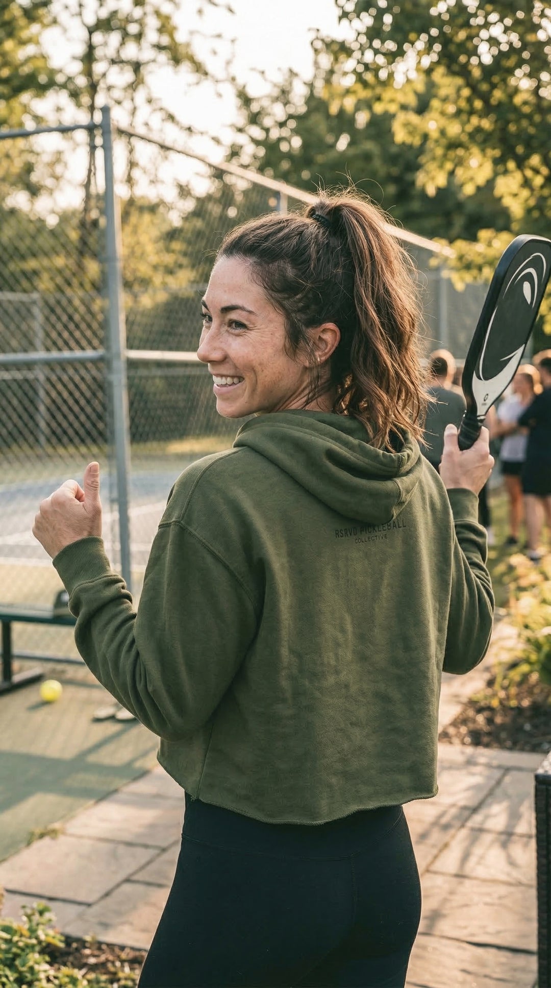 Woman in green hoodie holding a tennis racket on a court with trees in the background