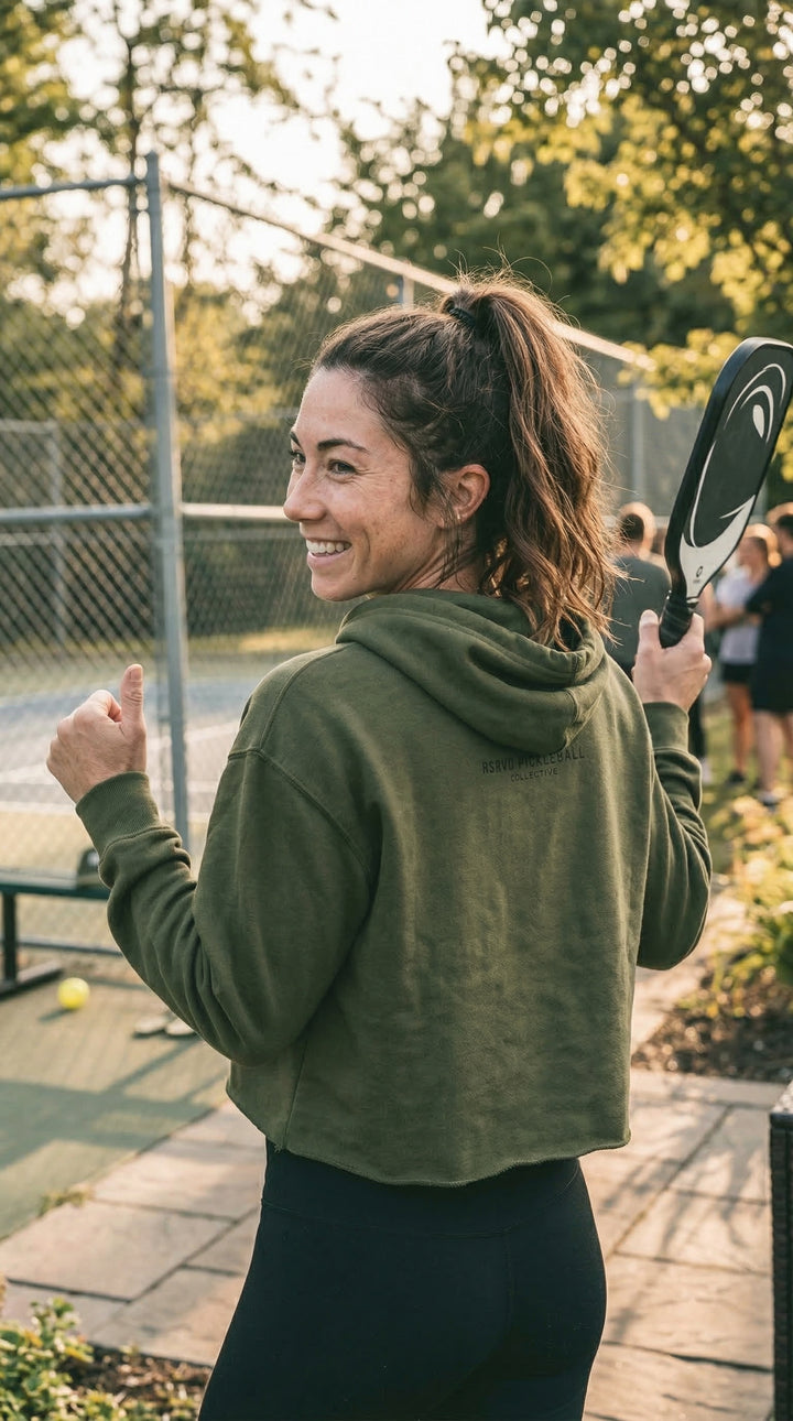 Woman in green hoodie holding a tennis racket on a court with trees in the background