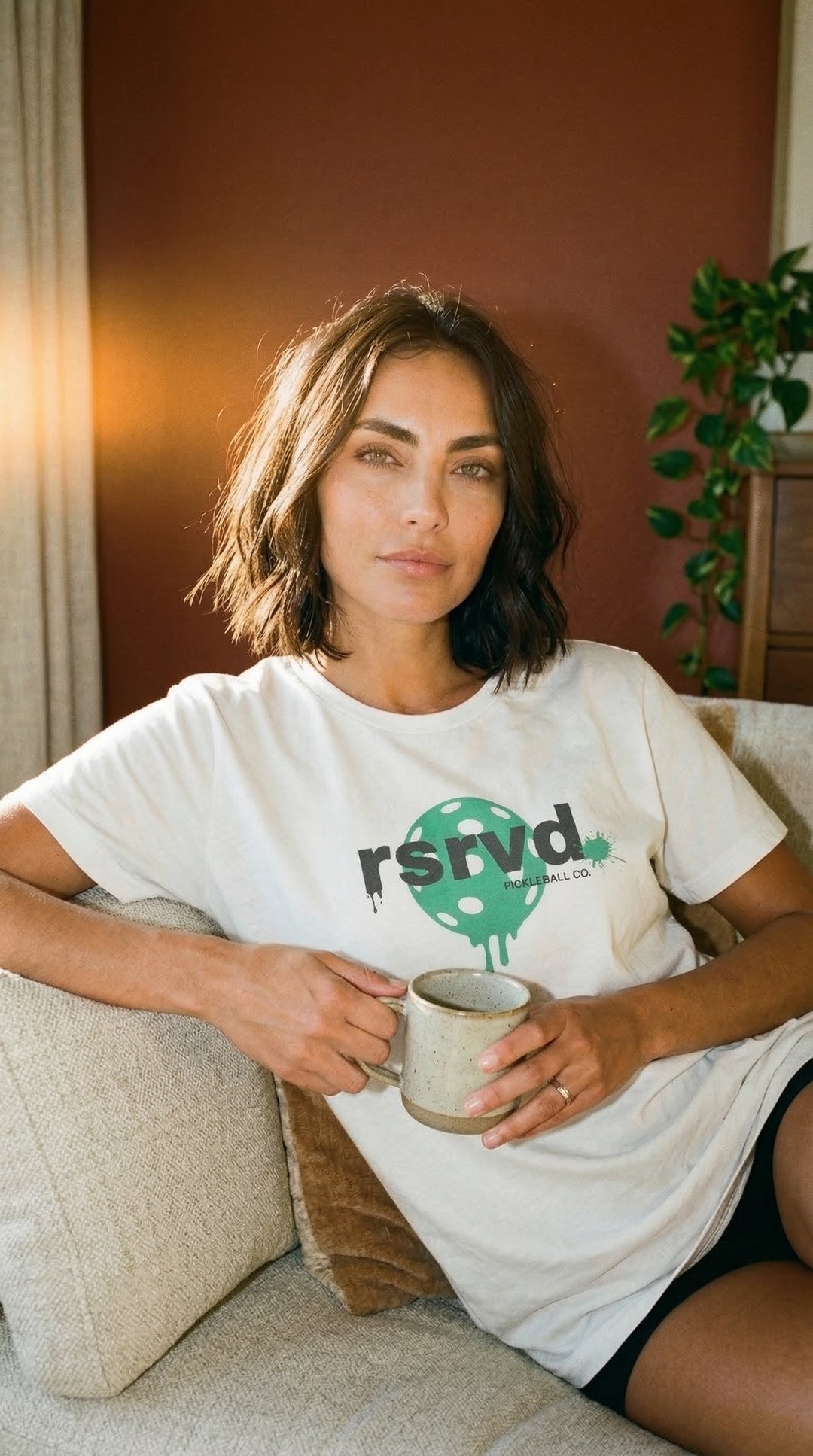 Woman sitting on a couch holding a mug, wearing a white t-shirt with a green logo and text.